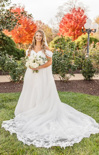 A bride in a white wedding gown holding a bouquet of white flowers, standing on the lawn of a historic estate with autumn foliage in the background.