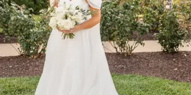 A bride in a white wedding gown holding a bouquet of white flowers, standing on the lawn of a historic estate with autumn foliage in the background.