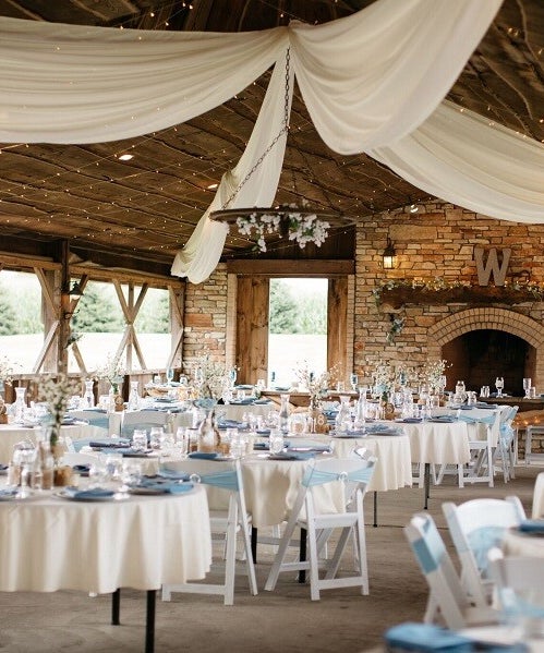 Interior of a rustic wedding venue pavilion with a stone fireplace, wooden beams, and tables set for a reception.