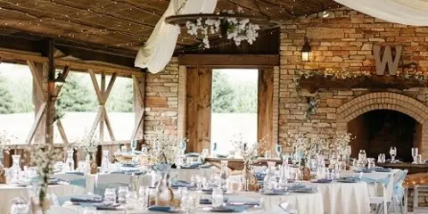 Interior of a rustic wedding venue pavilion with a stone fireplace, wooden beams, and tables set for a reception.
