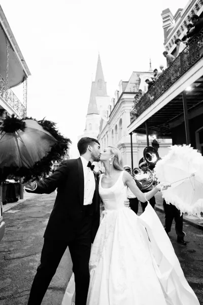 A black and white photograph of a bride and groom kissing on a street in New Orleans, surrounded by a brass band and onlookers on balconies.
