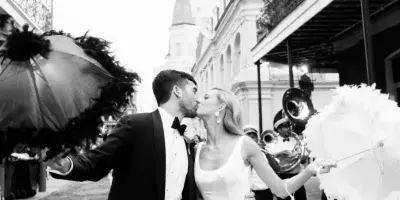 A black and white photograph of a bride and groom kissing on a street in New Orleans, surrounded by a brass band and onlookers on balconies.