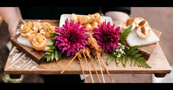 A wooden serving tray held by a person, displaying an assortment of gourmet appetizers including skewers and pastries, garnished with vibrant pink chrysanthemums and green ferns.