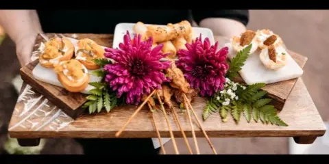 A wooden serving tray held by a person, displaying an assortment of gourmet appetizers including skewers and pastries, garnished with vibrant pink chrysanthemums and green ferns.