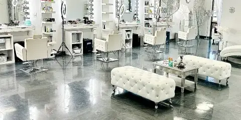 Interior view of a makeup studio featuring white chairs, vanity mirrors with lights, and product shelves.