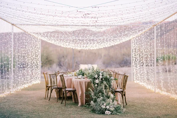 A wedding reception setup featuring a tent with string lights, tables with beige linens, wooden chairs, and a floral centerpiece.