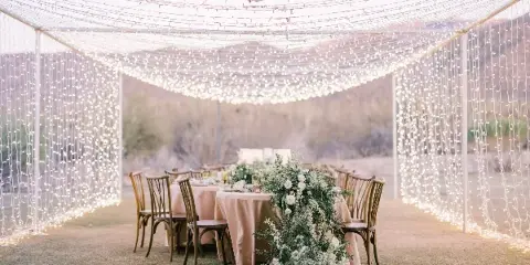A wedding reception setup featuring a tent with string lights, tables with beige linens, wooden chairs, and a floral centerpiece.