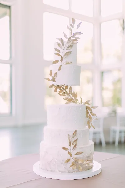 A five-tier white wedding cake with textured frosting and gold leaf decorations on a table.