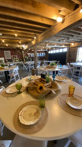 Interior view of a rustic barn venue featuring set round tables, white chairs, and exposed wooden ceiling beams.