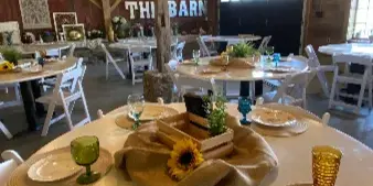 Interior view of a rustic barn venue featuring set round tables, white chairs, and exposed wooden ceiling beams.