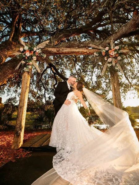 A bride and groom kissing under a rustic wooden arch decorated with flowers and pampas grass in an outdoor setting.