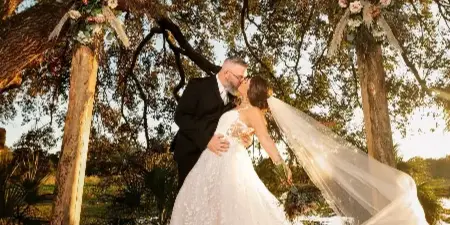 A bride and groom kissing under a rustic wooden arch decorated with flowers and pampas grass in an outdoor setting.