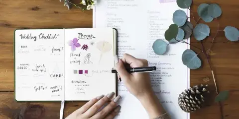 A person's hands writing in a wedding planner checklist and a notebook on a wooden table, surrounded by floral elements.