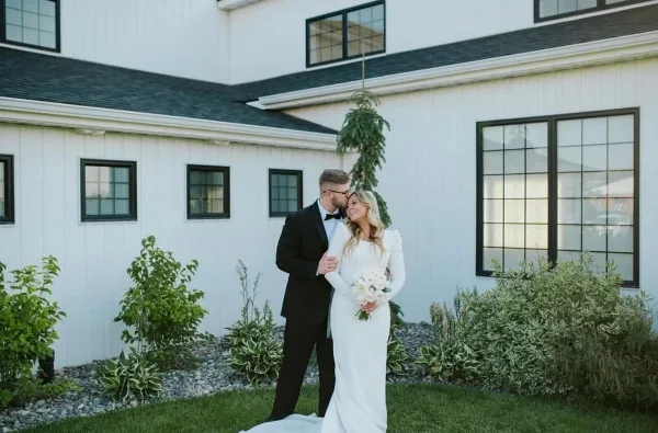 A bride and groom in formal wedding attire standing together outdoors in front of a white building with dark window frames.