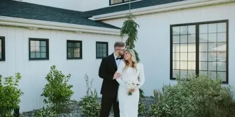 A bride and groom in formal wedding attire standing together outdoors in front of a white building with dark window frames.