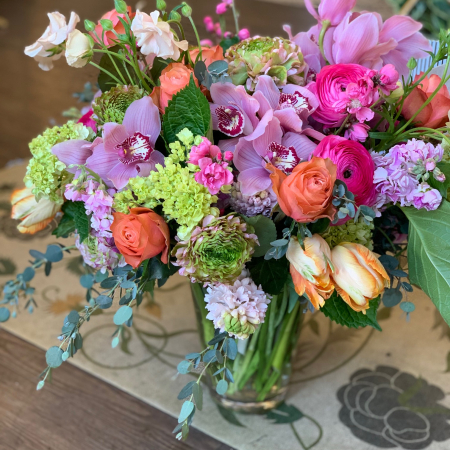 A colorful floral arrangement with pink orchids, orange roses, and green hydrangeas in a glass vase.