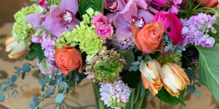 A colorful floral arrangement with pink orchids, orange roses, and green hydrangeas in a glass vase.