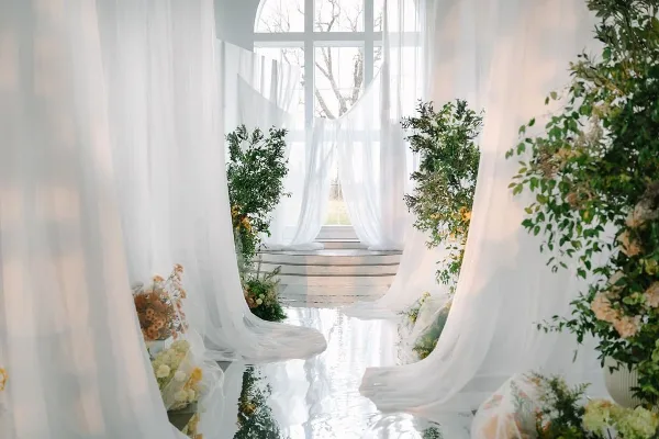 Wedding ceremony aisle decorated with white drapery, greenery, and floral arrangements in a bright venue.
