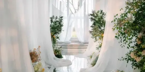 Wedding ceremony aisle decorated with white drapery, greenery, and floral arrangements in a bright venue.