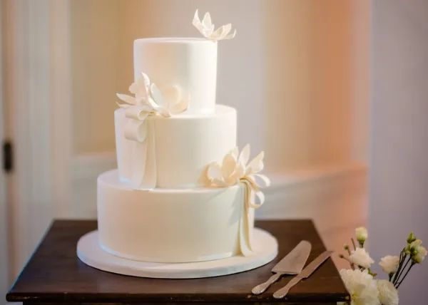 A three-tiered white wedding cake decorated with white sugar flowers and ribbons, sitting on a wooden table.