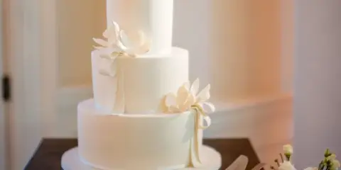 A three-tiered white wedding cake decorated with white sugar flowers and ribbons, sitting on a wooden table.