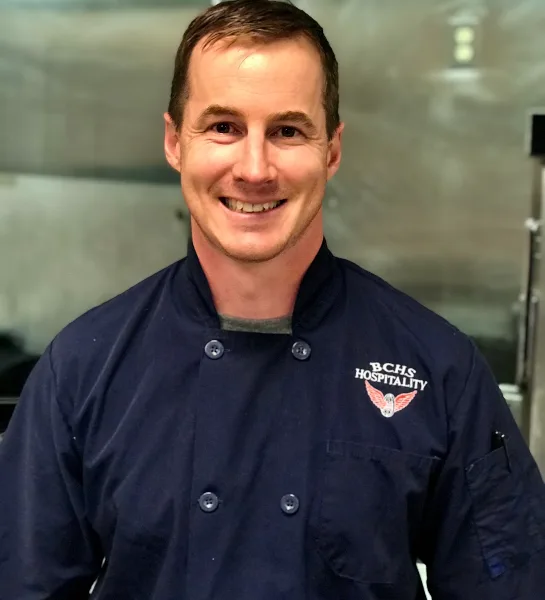 Portrait of a smiling chef in a dark blue chef's coat with 'BCHS HOSPITALITY' embroidered on the chest, standing in a commercial kitchen.
