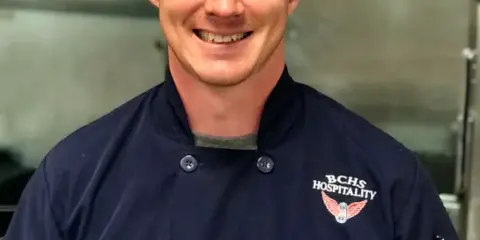 Portrait of a smiling chef in a dark blue chef's coat with 'BCHS HOSPITALITY' embroidered on the chest, standing in a commercial kitchen.