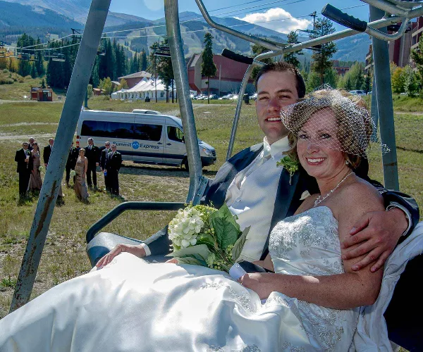 A bride and groom on a ski lift with a Summit Express shuttle van and wedding party visible in the background.