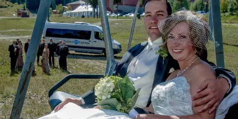 A bride and groom on a ski lift with a Summit Express shuttle van and wedding party visible in the background.