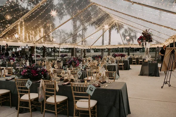 Interior of a large event tent with string lighting, starburst fixtures, and a formal dining setup with gold chiavari chairs and dark table linens.
