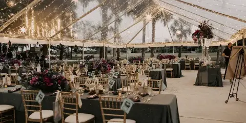 Interior of a large event tent with string lighting, starburst fixtures, and a formal dining setup with gold chiavari chairs and dark table linens.