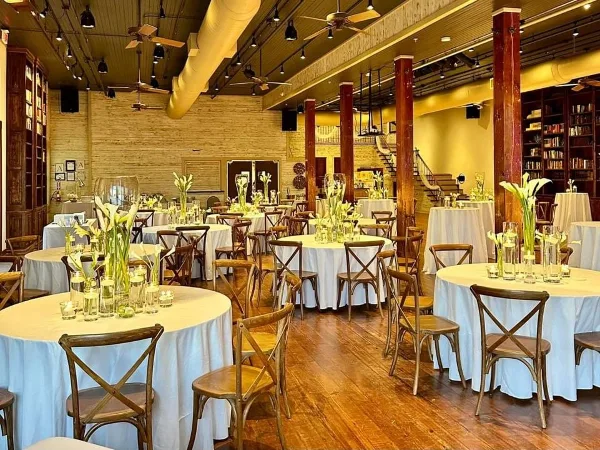 Interior of a wedding venue featuring round tables with white tablecloths, wooden chairs, and tall floral centerpieces in a room with exposed brick walls and wooden pillars.