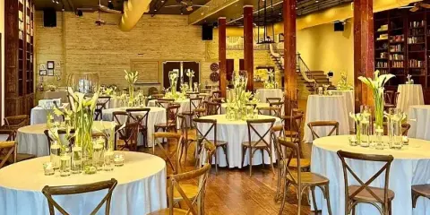 Interior of a wedding venue featuring round tables with white tablecloths, wooden chairs, and tall floral centerpieces in a room with exposed brick walls and wooden pillars.