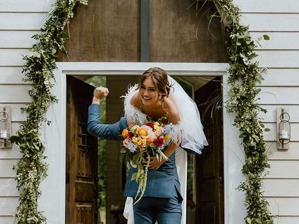 A bride and groom standing at the doorway of a white cottage decorated with greenery.