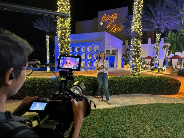 A videographer filming a woman standing outdoors at night in front of a building with palm trees and string lights.