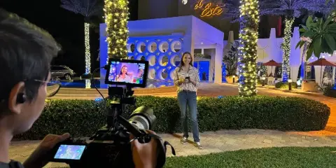 A videographer filming a woman standing outdoors at night in front of a building with palm trees and string lights.