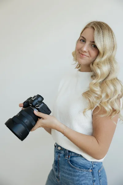 A blonde woman in a white top and jeans holding a professional DSLR camera.