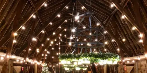 Interior view of a rustic barn venue featuring exposed wooden beams, string lights, and a large decorative chandelier.