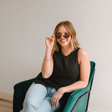 Portrait of a smiling woman with blonde hair wearing sunglasses, a black sleeveless top, and jeans, sitting in a green chair.
