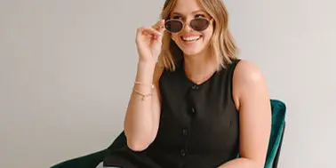 Portrait of a smiling woman with blonde hair wearing sunglasses, a black sleeveless top, and jeans, sitting in a green chair.