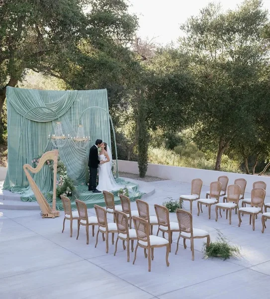 Outdoor wedding ceremony setup with a green draped altar, chandelier, harp, and rows of chairs.