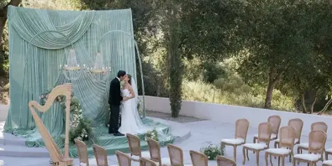 Outdoor wedding ceremony setup with a green draped altar, chandelier, harp, and rows of chairs.