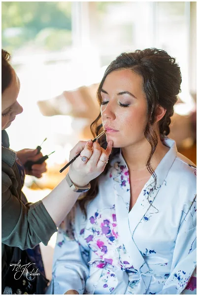 A makeup artist applying lip color to a bride wearing a floral robe.