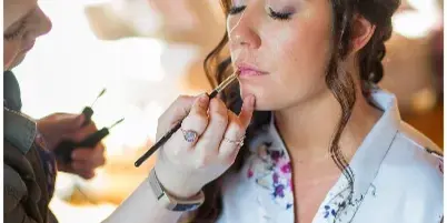 A makeup artist applying lip color to a bride wearing a floral robe.