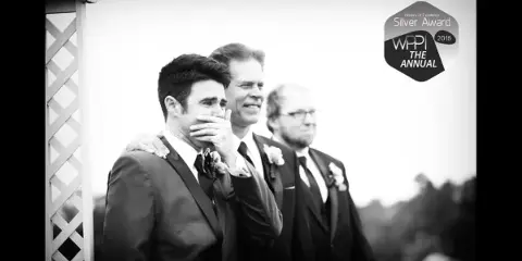 A black and white photograph of a groom covering his mouth with his hand, standing with his groomsmen during a wedding ceremony.