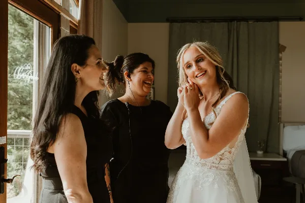 A wedding coordinator wearing a headset microphone smiles while assisting a bride in a white lace wedding dress.