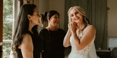 A wedding coordinator wearing a headset microphone smiles while assisting a bride in a white lace wedding dress.
