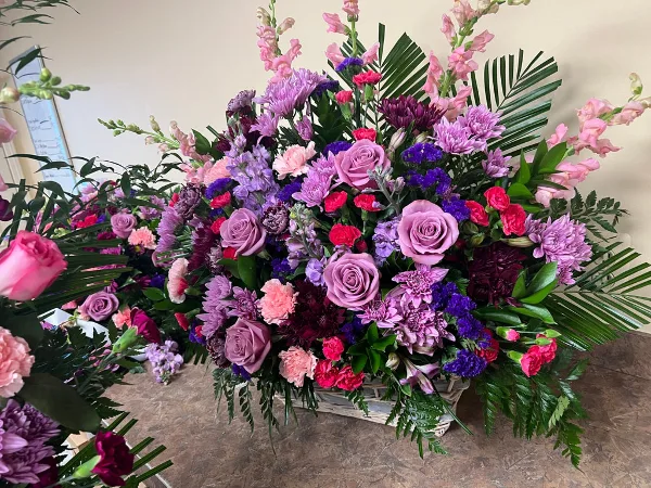 A large, colorful floral arrangement featuring pink roses, purple chrysanthemums, and greenery in a decorative basket.