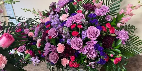 A large, colorful floral arrangement featuring pink roses, purple chrysanthemums, and greenery in a decorative basket.