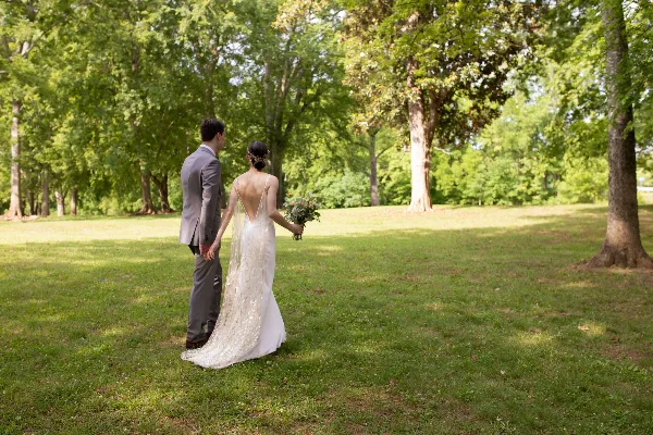 A bride and groom standing in a grassy field with trees in the background, holding hands and a bouquet.